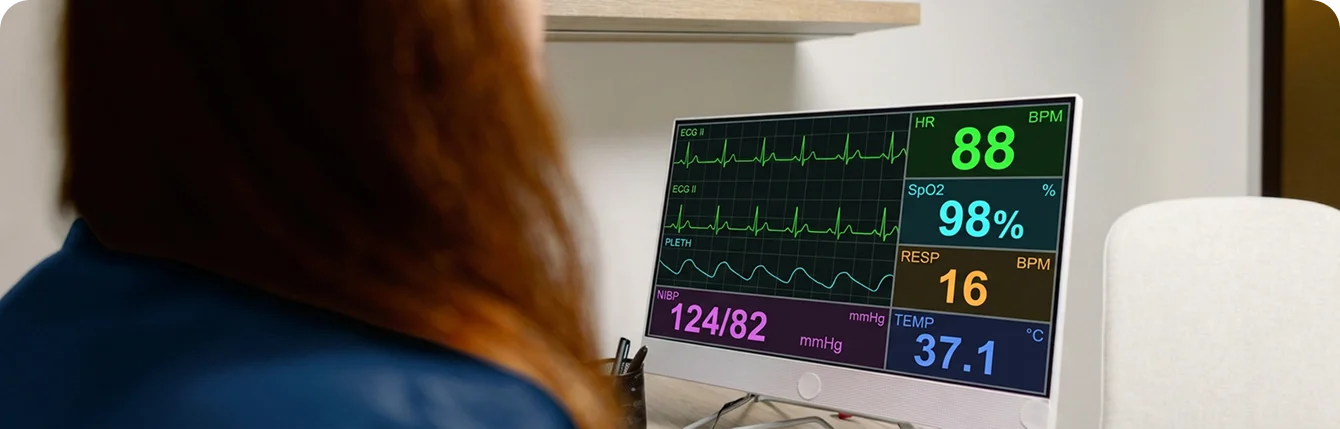 Healthcare professional observing a bedside cardiac monitor displaying ECG waveforms, heart rate, oxygen saturation, blood pressure, and patient vital signs.