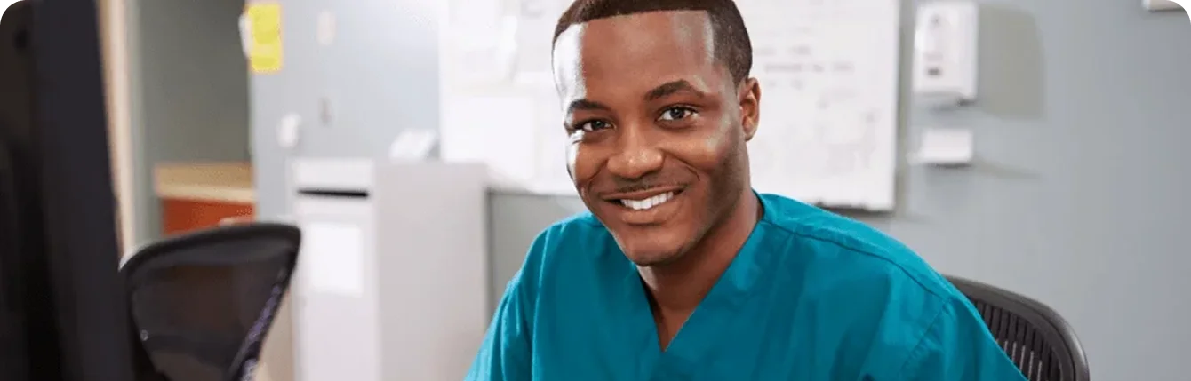 Smiling healthcare professional wearing teal medical scrubs at a clinical workstation, representing healthcare staffing and virtual nursing partnerships.