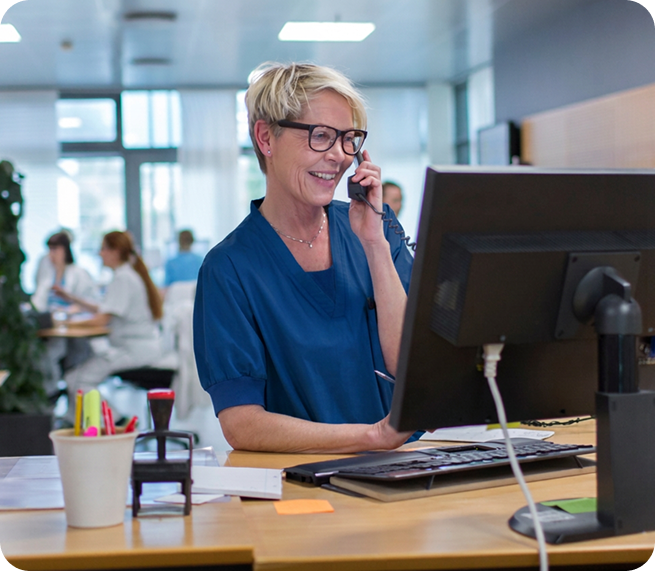 Healthcare professional wearing blue scrubs and glasses, speaking on a headset while working at a computer to coordinate clinical care services.